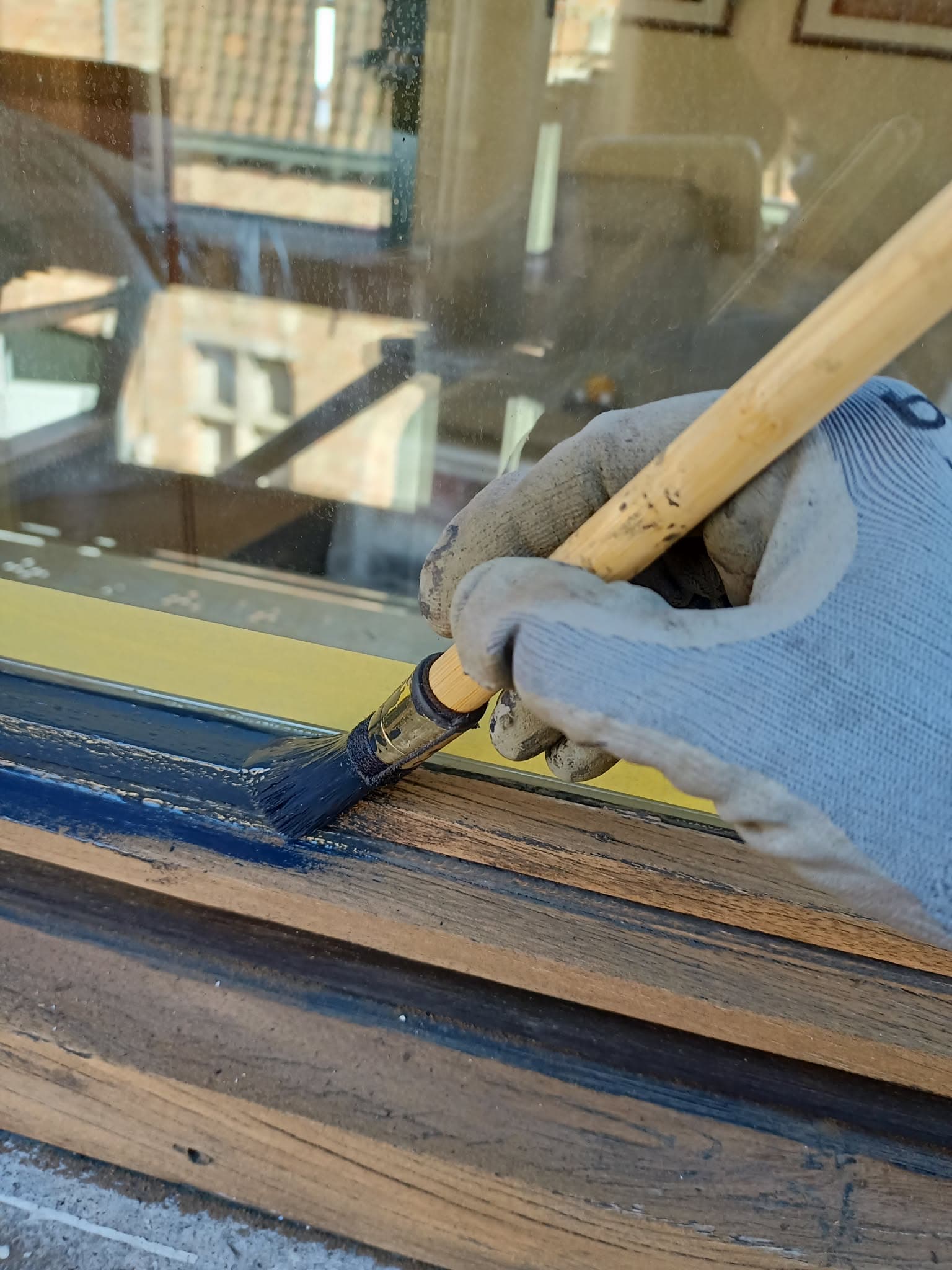 Main gantée peignant un cadre de fenêtre en bois avec de la peinture bleu foncé.